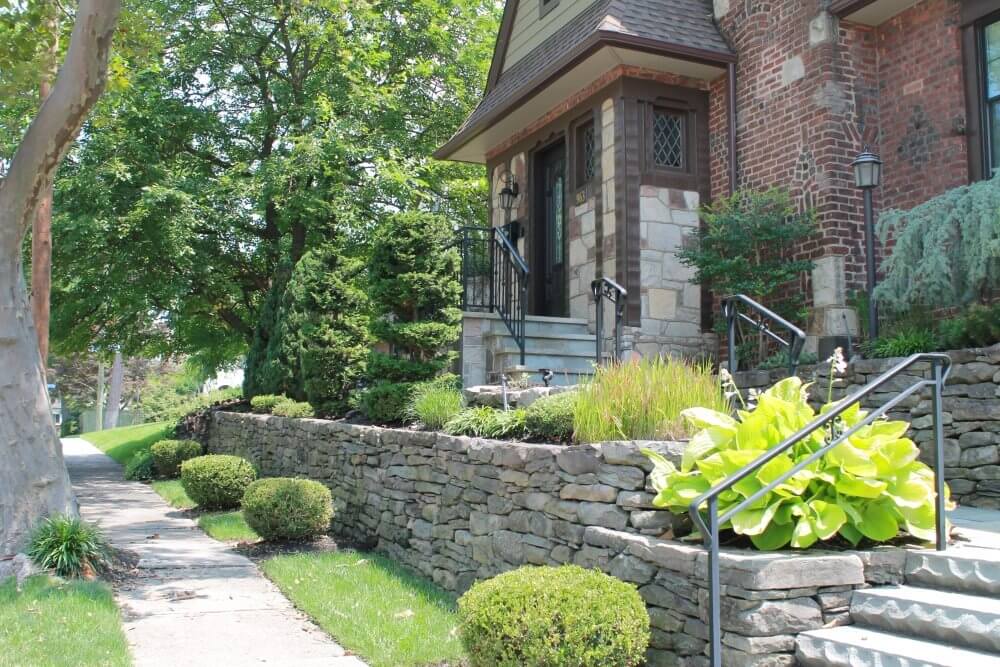 A stone pathway and stairs lead up to the front door of a brick house surrounded by greenery and well-manicured bushes. A tree and sidewalk are visible on the left side of the image.