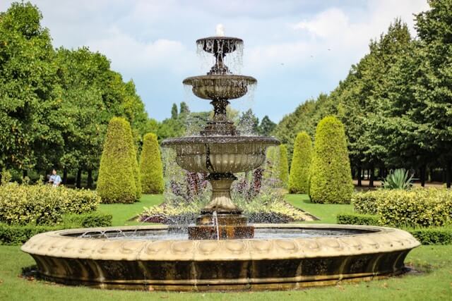 An ornate three-tiered fountain in a lush garden with manicured bushes and trees under a clear sky.