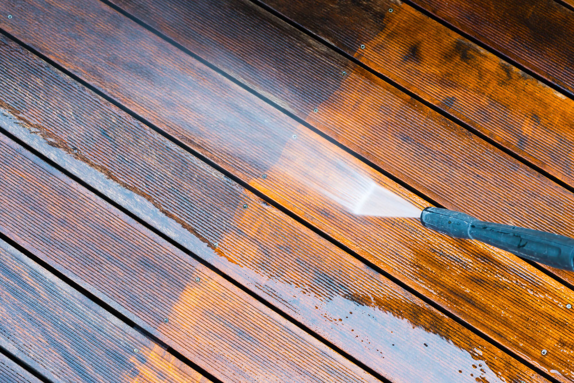 A power washer cleaning a deck with wooden planks, showing a stark contrast between the dirty and cleaned areas.